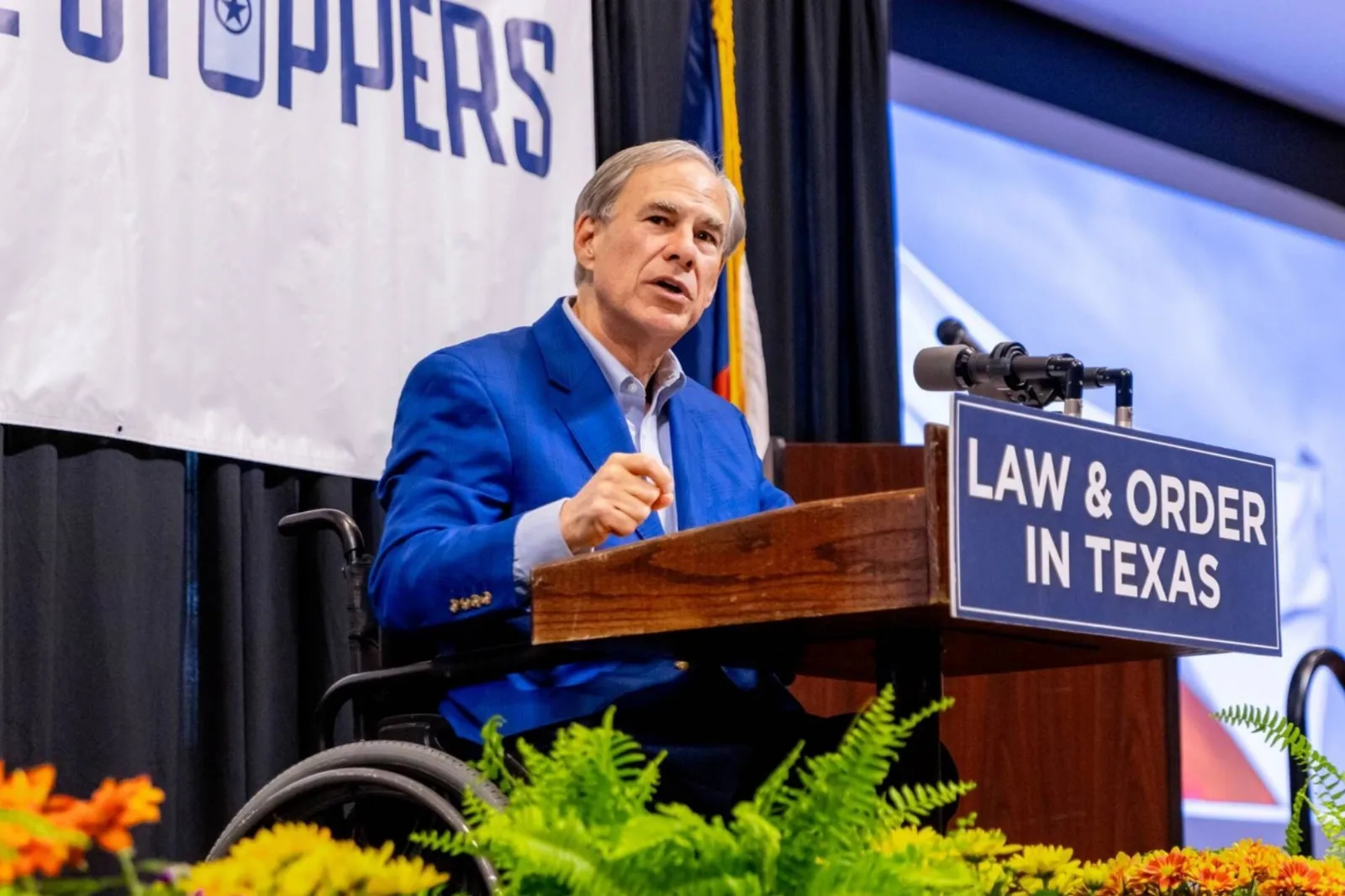 Greg Abbott en conferencia de prensa con fondo de bandera de Texas y hospital al fondo