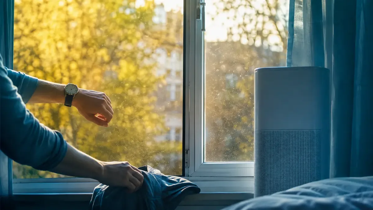 Ventana entreabierta con polen afuera mientras una mano revisa la hora y retira ropa del alféizar