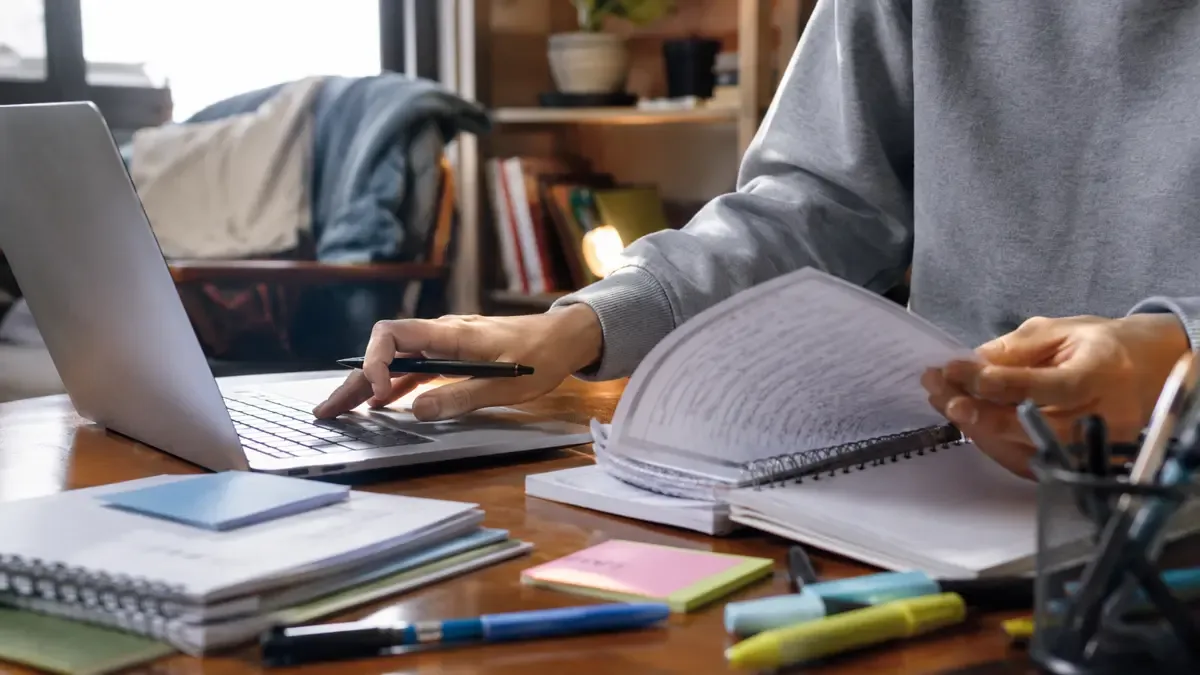 Manos adultas organizando apuntes y una notebook en un rincón doméstico de estudio online, vista en tres cuartos, profundidad de campo suave
