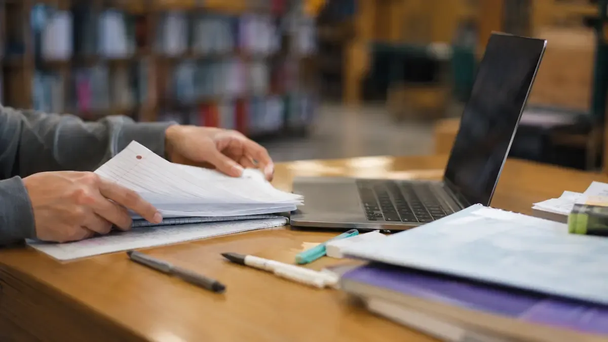 Manos adultas organizando apuntes y una notebook en una mesa de biblioteca, plano medio documental, perspectiva natural